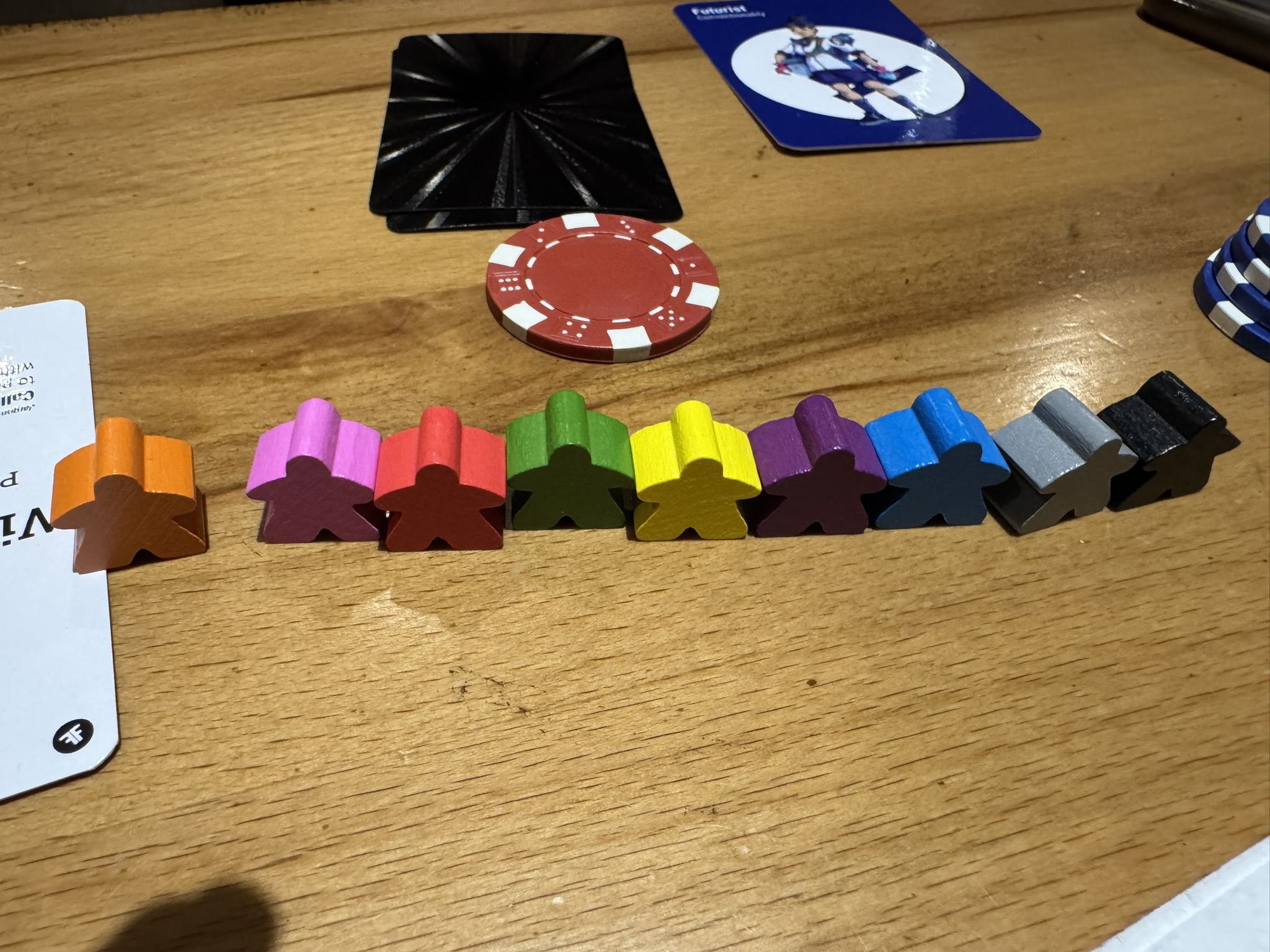 Colourful wooden meeple player tokens lined up on a Game of Gains table alongside poker chips and cards
